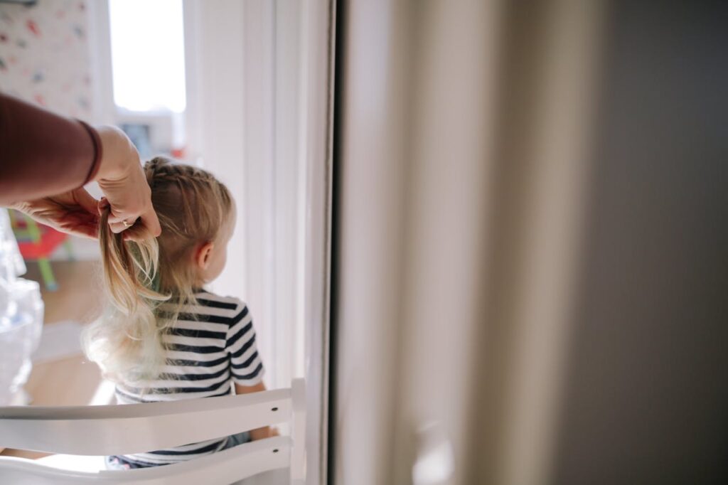 A mother gently braids her daughter's hair inside a sunlit room, capturing a tender family moment.